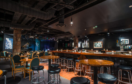A darkly lit nightclub with bar stool tables, sit down tables and stage with piano and drum set.