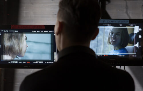 A director watching one of two TV screens in front of him. Each screen showing a different camera angle of the same woman.