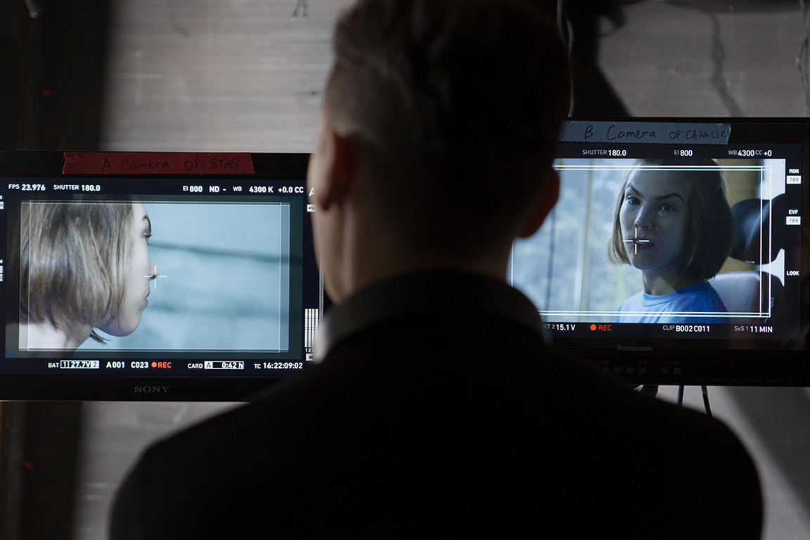 A director watching one of two TV screens in front of him. Each screen showing a different camera angle of the same woman.