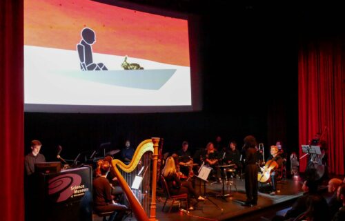 A theater stage with a group of musicians. A video is being projected behind them