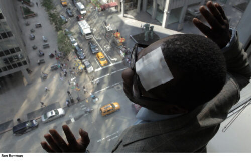 A man with a bandage on his forehead looking out a window of a tall building with his hands raised up. Pedestrians and cars on the street can be seen below.