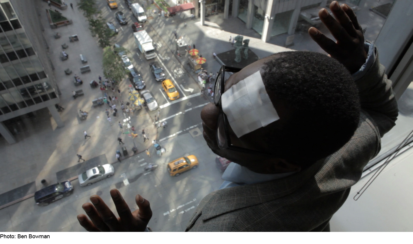 A man with a bandage on his forehead looking out a window of a tall building with his hands raised up. Pedestrians and cars on the street can be seen below.