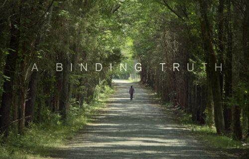 Title text "A Binding Truth". A man walking down a dirt path parted through tall trees on each side.
