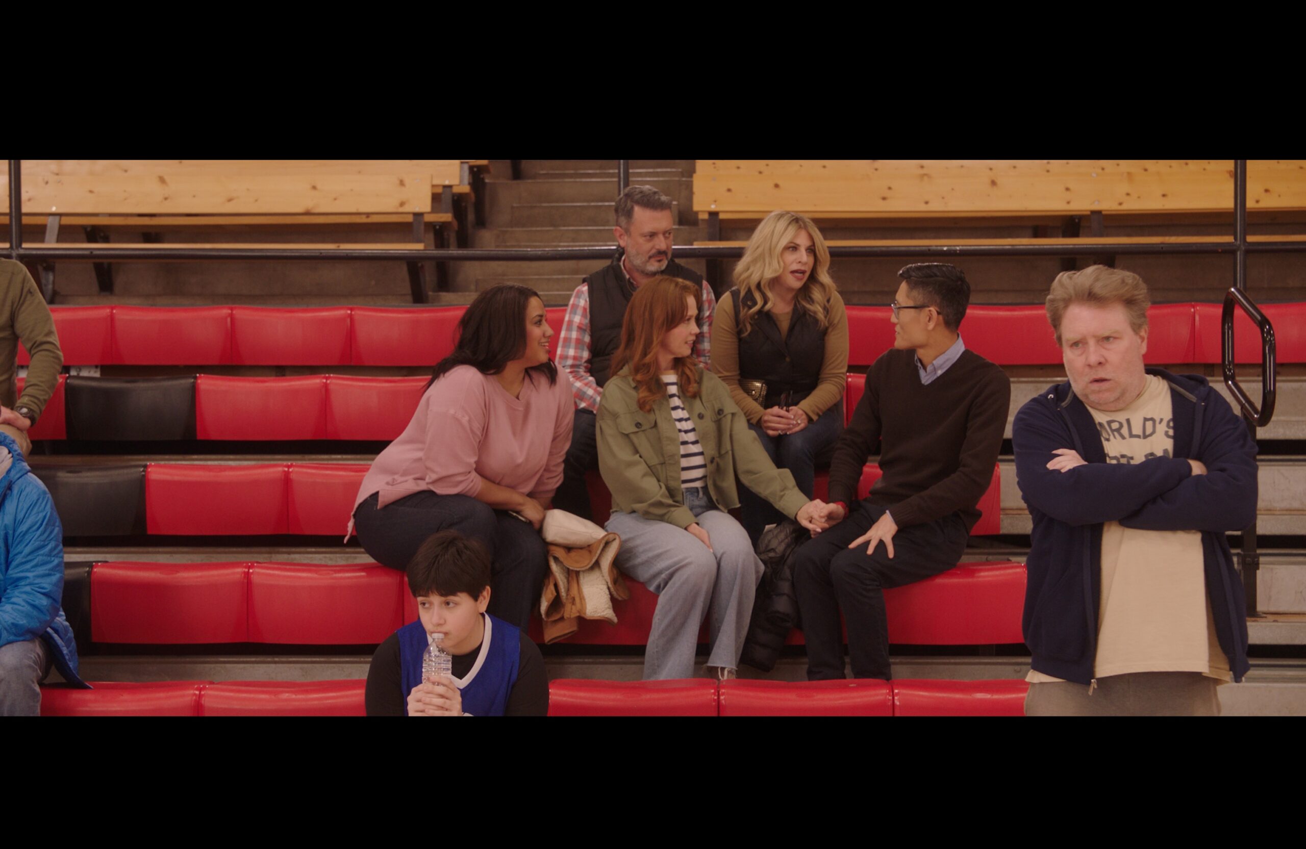 A group of people sitting in auditorium stands while a coach stands in the foreground with his arms crossed